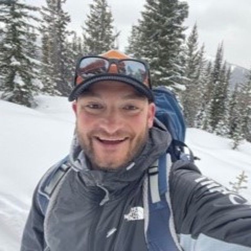 The image shows a man in a snowy, mountainous environment, likely hiking or engaging in a winter activity. He is wearing a jacket, a backpack, and a cap, and sunglasses. The background features snow-covered trees and mountains, suggesting a cold climate. The man is smiling, indicating he is enjoying the outdoor experience.
